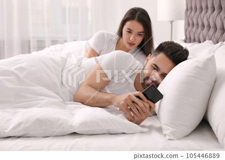 Distrustful young woman peering into boyfriend's smartphone in bed at home 105446889