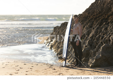 A smiling man with a naked torso standing by a rock holding a surfboard 105448550