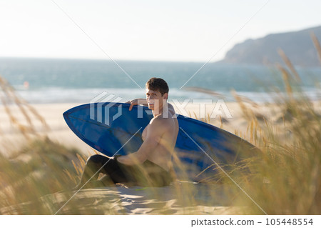 A man sitting on the beach holding a blue surfboard - looking in the camera A man sitting on the beach holding a blue surfboard - looking in the camera 105448554