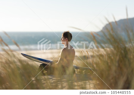 A man sitting on the beach holding a blue surfboard 105448555