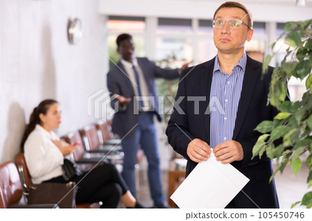 Upset adult man in glasses with documents standing in reception 105450746