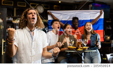 Cheerful international soccer football fans waving Russian flag and drinking beer, eating chips in the sport bar Cheerful international soccer football fans waving Russian flag and drinking beer, eating chips in the sport bar 105450914