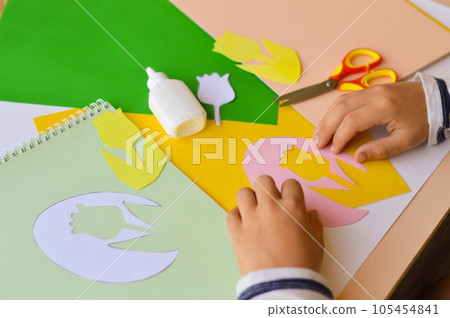 A small child makes paper crafts for mother's day or birthday. A boy makes paper flowers for mom. A simple pleasant idea of a children's gift to mom. 105454841