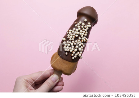 Human Hand Holds Penis Shaped Waffle On Wooden Stick With Dark Chocolate, Decorated With White Air Corn Balls On Pink Background. Festival Street Food, Sweet Cookie Horizontal Plane. Copy Space 105456355