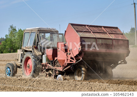 Spring sowing on farmer field, an agricultural wheeled tractor with hitched potato planter rides on arable land. 105462143