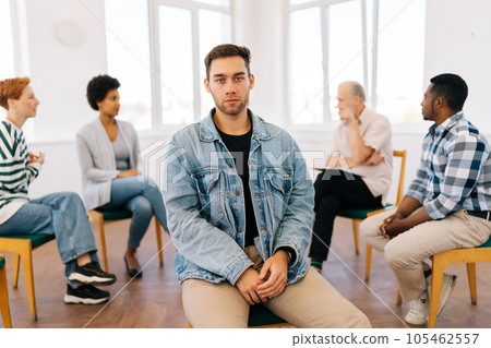 Portrait of sad Caucasian man looking at camera with serious expression while group of multicultural and different ages people sitting in circle, talking in background during therapy session. Portrait of sad Caucasian man looking at camera with serious expression while group of multicultural and different ages people sitting in circle, talking in background during therapy session. 105462557