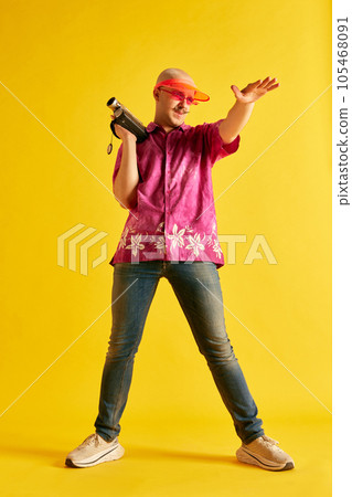 Image of full lengh young man in bright cap and t-shirt reguleting process of shooting on vintage camera against bright yellow background. Image of full lengh young man in bright cap and t-shirt reguleting process of shooting on vintage camera against bright yellow background. 105468091