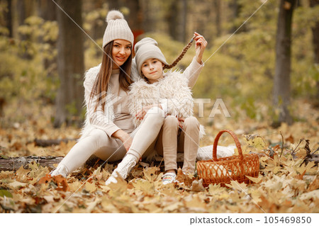 Young woman with little girl sitting on a fallen tree trunk in autumn forest. Brunette woman play with her daughter. Girl wearing beige sweater and mother wearing white clothes. 105469850