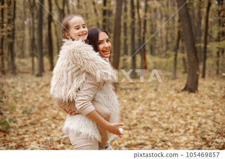 Young woman with little girl standing in autumn forest. Brunette woman holding her daughter piggyback. Girl wearing beige sweater and mother wearing white clothes. 105469857