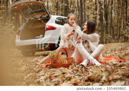 Young woman with little girl sitting on a red blanket in autumn forest. Brunette woman play with her daughter. Girl wearing beige sweater and mother wearing white clothes. Young woman with little girl sitting on a red blanket in autumn forest. Brunette woman play with her daughter. Girl wearing beige sweater and mother wearing white clothes. 105469862