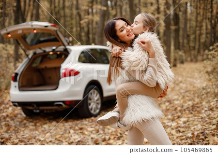 Young woman with little girl standing in autumn forest near their car. Brunette woman play with her daughter. Girl wearing beige sweater and mother wearing white clothes. 105469865