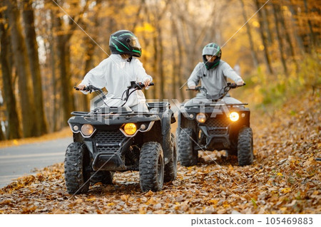 A young caucasian man and woman riding an ATV quad bikes in autumn forest. Two friends maneuvering off-road ATV. Couple wearing grey sportive costumes. A young caucasian man and woman riding an ATV quad bikes in autumn forest. Two friends maneuvering off-road ATV. Couple wearing grey sportive costumes. 105469883