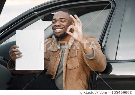 Black man holding blank sheet of paper in his hand. Man sitting in a car and wearing brown jacket. Man has happy face impression and showing sign okay. Black man holding blank sheet of paper in his hand. Man sitting in a car and wearing brown jacket. Man has happy face impression and showing sign okay. 105469908