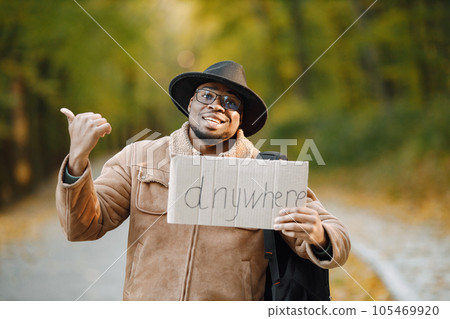 Young black man hitchhiking on road and holding a sign anywhere. Male traveler traveling alone by autostop showing thumb up gesture outdoors. Man wearing brown jacket, black hat and backpack. 105469920
