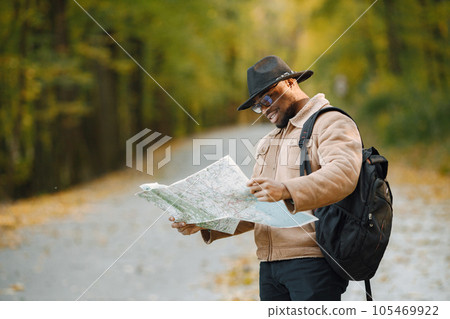Young black man hitchhiking on road and looking at map. Male traveler feeling lost, traveling alone by autostop. Man wearing brown coat, black hat and backpack. 105469922
