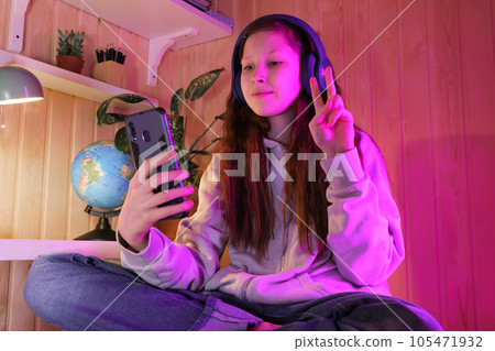 Happy cute pretty teen in modern headphones joying, in neon room at home. Girl making selfie, listen favorite music, making video call. Have fun alone at home during lockdown 105471932
