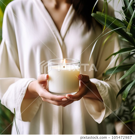 Young woman holding burning candle jar in her hands, container candle mockup closeup shot, mindfulness home interior with green plants 105472987