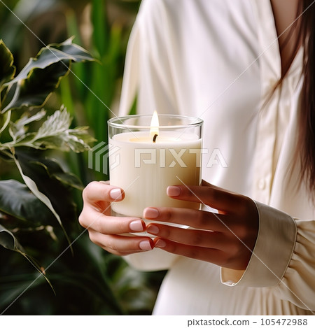 Young woman holding burning candle jar in her hands, container candle mockup closeup shot, mindfulness home interior with green plants 105472988