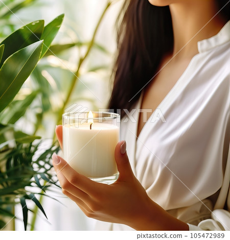 Young woman holding burning candle jar in her hands, container candle mockup closeup shot, mindfulness home interior with green plants 105472989