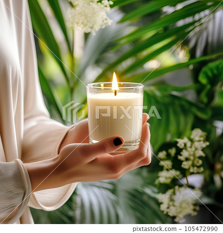Young woman holding burning candle jar in her hands, container candle mockup closeup shot, mindfulness home interior with green plants 105472990