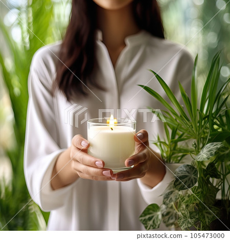 Young woman holding burning candle jar in her hands, container candle mockup closeup shot, mindfulness home interior with green plants 105473000