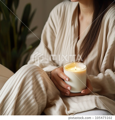 Beautiful young woman with long hair dressed in white relaxing in her home bedroom with burning candles, flowers and books, cozy home decor in natural colors. Candle jar mockup 105473102