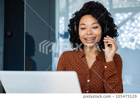 Young beautiful woman talking to customers remotely, african american female worker smiling using headset phone and laptop for online consultation, customer service. Young beautiful woman talking to customers remotely, african american female worker smiling using headset phone and laptop for online consultation, customer service. 105473129