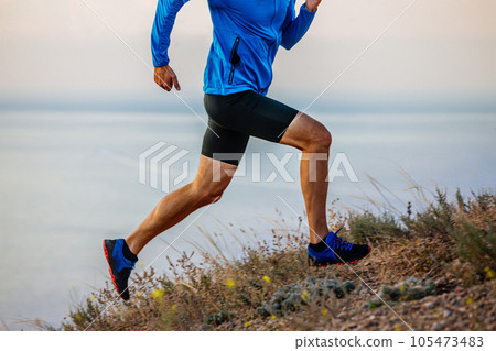 close-up male runner run uphill on trail in blue jacket and black tights, background of sky and sea 105473483