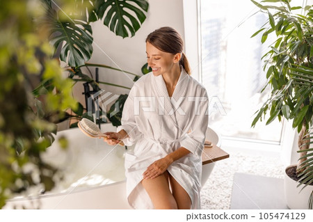 Cute young woman in bathrobe smiling while holding natural brush, sitting on the side of a bathtub in the bathroom decorated with green plants Cute young woman in bathrobe smiling while holding natural brush, sitting on the side of a bathtub in the bathroom decorated with green plants 105474129
