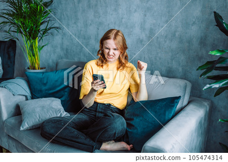 Confused angry woman having problem with phone, sitting on couch at home, unhappy young female looking at screen, dissatisfied by discharged or broken smartphone, reading bad news in message Confused angry woman having problem with phone, sitting on couch at home, unhappy young female looking at screen, dissatisfied by discharged or broken smartphone, reading bad news in message 105474314