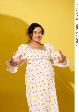 Multi-ethnic happy pregnant woman points fingers at her big belly in late pregnancy in 38 week, smiles looking at camera 105475187