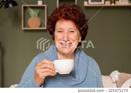 Portrait of an elderly woman with blue eyes looks at the camera and laughs with a cup of coffee in her hands Portrait of an elderly woman with blue eyes looks at the camera and laughs with a cup of coffee in her hands 105475274