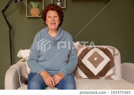 Portrait of a modern elderly woman with blue eyes and red hair sits on a sofa and looks at the camera Portrait of a modern elderly woman with blue eyes and red hair sits on a sofa and looks at the camera 105475465