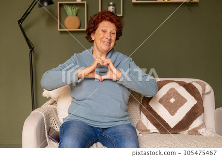 Portrait of a modern elderly woman with blue eyes and red hair sits on a sofa, smiles and shows a heart gesture with her hands Portrait of a modern elderly woman with blue eyes and red hair sits on a sofa, smiles and shows a heart gesture with her hands 105475467