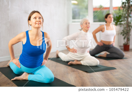 Smiling women practicing pigeon pose of yoga in light fitness room Smiling women practicing pigeon pose of yoga in light fitness room 105479422