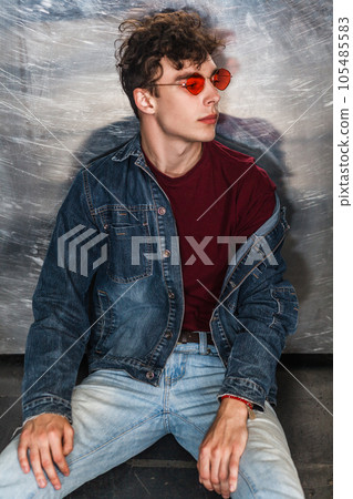 Portrait of trendy stylish attractive fashion model man in bright red sunglasses and denim casual style sitting on floor near metallic wall and looking away. Indoor studio shot. 105485583