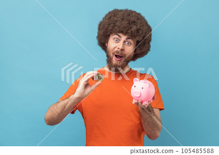 Portrait of man with Afro hairstyle wearing orange T-shirt holding piggy bank and golden bitcoin, looking at camera with shocked expression. Indoor studio shot isolated on blue background. Portrait of man with Afro hairstyle wearing orange T-shirt holding piggy bank and golden bitcoin, looking at camera with shocked expression. Indoor studio shot isolated on blue background. 105485588