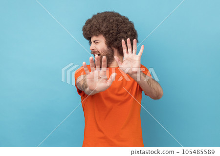 Portrait of scared man with Afro hairstyle wearing orange T-shirt standing with afraid or worry face, looking at camera and blocking with his hands. Indoor studio shot isolated on blue background. Portrait of scared man with Afro hairstyle wearing orange T-shirt standing with afraid or worry face, looking at camera and blocking with his hands. Indoor studio shot isolated on blue background. 105485589