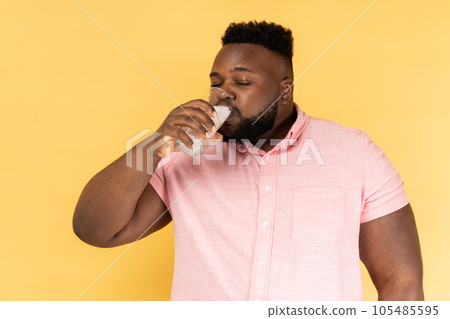 Portrait of bearded man wearing pink shirt drinking water with ice from glass, feels thirsty, enjoying cold beverage during hot summer days. Indoor studio shot isolated on yellow background. Portrait of bearded man wearing pink shirt drinking water with ice from glass, feels thirsty, enjoying cold beverage during hot summer days. Indoor studio shot isolated on yellow background. 105485595
