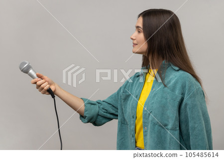 Side view of satisfied woman with dark hair standing offering microphone, journalist or reporter taking interview, wearing casual style jacket. Indoor studio shot isolated on gray background. 105485614