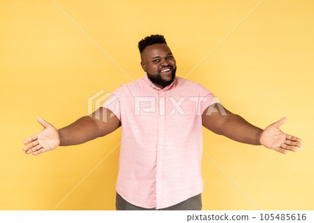 Portrait of happy generous bearded man wearing pink shirt standing with raised hands and looking at camera, welcoming or giving smth. Indoor studio shot isolated on yellow background. Portrait of happy generous bearded man wearing pink shirt standing with raised hands and looking at camera, welcoming or giving smth. Indoor studio shot isolated on yellow background. 105485616
