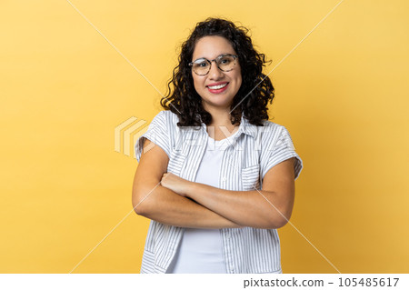 Portrait of charming delighted happy woman in optical eyeglasses with dark wavy hair standing with folded hands, smiling to camera. Indoor studio shot isolated on yellow background. Portrait of charming delighted happy woman in optical eyeglasses with dark wavy hair standing with folded hands, smiling to camera. Indoor studio shot isolated on yellow background. 105485617