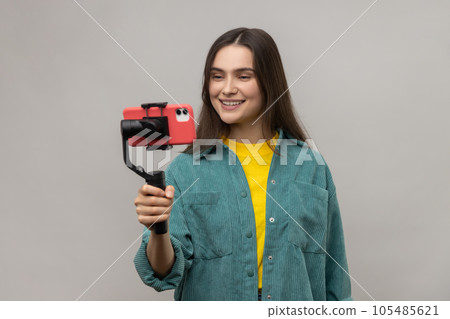 Trendy beautiful woman holding smartphone on steadycam in hand, blogger making selfie for social network, live stream, wearing casual style jacket. Indoor studio shot isolated on gray background. Trendy beautiful woman holding smartphone on steadycam in hand, blogger making selfie for social network, live stream, wearing casual style jacket. Indoor studio shot isolated on gray background. 105485621