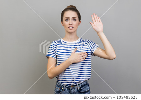Portrait of serious young adult woman wearing striped T-shirt holding on chest another, swearing in honesty and devotion, patriotism. Indoor studio shot isolated on gray background. Portrait of serious young adult woman wearing striped T-shirt holding on chest another, swearing in honesty and devotion, patriotism. Indoor studio shot isolated on gray background. 105485623