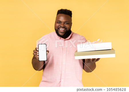 Portrait of positive man wearing pink shirt holding present box and smatrphone with blank screen for promotion, looking smiling at camera. Indoor studio shot isolated on yellow background. 105485628
