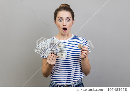 Portrait of surprised shocked woman wearing striped T-shirt holding gold bitcoin and fan of dollar bills, looking at camera with open mouth. Indoor studio shot isolated on gray background. Portrait of surprised shocked woman wearing striped T-shirt holding gold bitcoin and fan of dollar bills, looking at camera with open mouth. Indoor studio shot isolated on gray background. 105485658