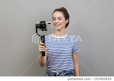 Portrait of smiling satisfied woman blogger wearing striped T-shirt using steadicam and cell phone for livestream, talking to followers. Indoor studio shot isolated on gray background. Portrait of smiling satisfied woman blogger wearing striped T-shirt using steadicam and cell phone for livestream, talking to followers. Indoor studio shot isolated on gray background. 105485659