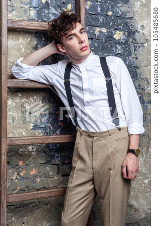 Portrait of handsome serious man in white shirt and beige pants leans on old wooden ladder, looking at camera, waiting for something, posing on brick wall background. Indoor studio shot. Portrait of handsome serious man in white shirt and beige pants leans on old wooden ladder, looking at camera, waiting for something, posing on brick wall background. Indoor studio shot. 105485680