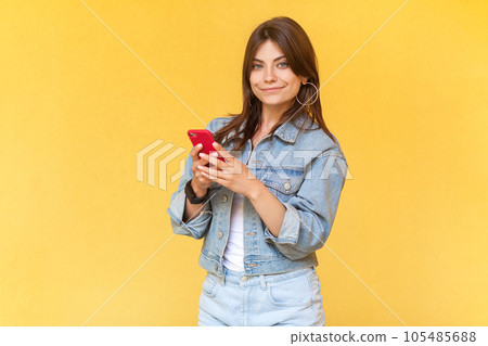 Portrait of adorable good looking young adult woman in denim jacket standing with smart phone in hands, surfing Internet, checking social networks. Indoor studio shot isolated on yellow background. Portrait of adorable good looking young adult woman in denim jacket standing with smart phone in hands, surfing Internet, checking social networks. Indoor studio shot isolated on yellow background. 105485688