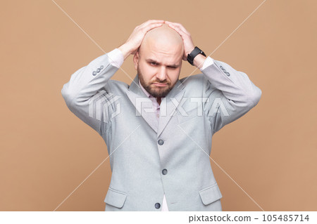 Depressed ill bald bearded man frowning and clasping sore head, suffering intense headache, having unbearable migraine, wearing gray jacket. Indoor studio shot isolated on brown background. Depressed ill bald bearded man frowning and clasping sore head, suffering intense headache, having unbearable migraine, wearing gray jacket. Indoor studio shot isolated on brown background. 105485714